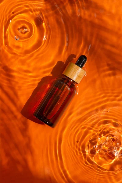 Amber bottle with dropper pipette with serum or essential oil on beautiful bursts and glare. Skincare cosmetic on orange background. Natural sunlight and water spills. Summer mood.