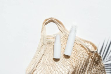 Krem ya da losyon için plastik beyaz tüpler ve bez alışveriş çantası. Beyaz arka planda güneş kremi ve palmiye yapraklarından gölgeler. Yüz bakımı için güzel yaz konsepti