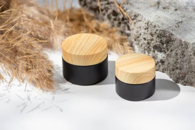 Kozmetik siyah kavanoz, kremalı ahşap kapak, lotyon ve kuru bitki. Cilt bakım ürünleri ve kır çiçekleri. Beyaz arka plan, gün ışığı ve güzel gölgeler. Yüz ve vücut bakımı için güzellik konsepti