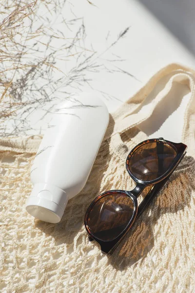 Plastic white tube for cream or lotion with sunglasses and loth shopping bag. Sunscreen cosmetic on white background with beautiful shadows and field grass. Beauty summer concept for face care