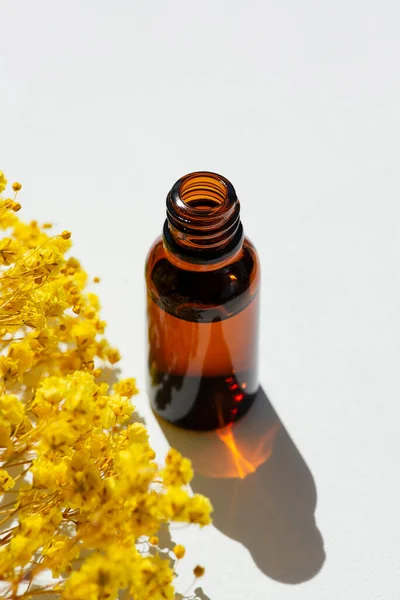 Open amber bottle without cap with serum or essential oil with beautiful yellow flowers. White background with daylight and beautiful shadows. Beauty concept for face and body care