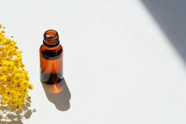 Open amber bottle without cap with serum or essential oil with yellow flowers. White background with daylight and beautiful shadows. Beauty concept for face and body care