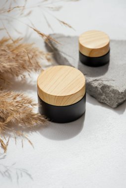 Kozmetik siyah kavanoz, kremalı ahşap kapak, lotyon ve kuru bitki. Cilt bakım ürünleri ve kır çiçekleri. Beyaz arka plan, gün ışığı ve güzel gölgeler. Yüz ve vücut bakımı için güzellik konsepti