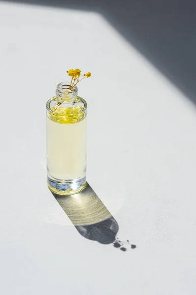 Open transparent bottle without cap with serum or essential oil with beautiful yellow flowers in soft focus. White background with daylight and beautiful shadows. Beauty concept for face and body care