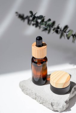 Kehribar şişe damlası pipeti ve serum ya da gerekli yağ, kozmetik siyah kavanoz ahşap krem ya da okaliptüs dallı gri beton podyumda lotyon. Beyaz arka plan, gün ışığı. Yüz ve vücut bakımı için güzellik konsepti