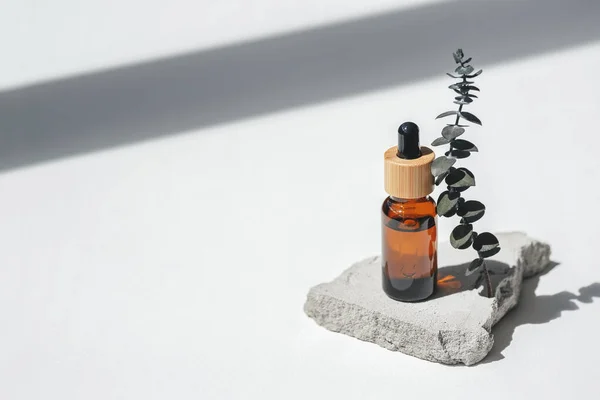 Amber bottle with dropper pipette and serum or essential oil on grey concrete podium with eucalyptus branches. White background with daylight. Beauty concept for face and body care
