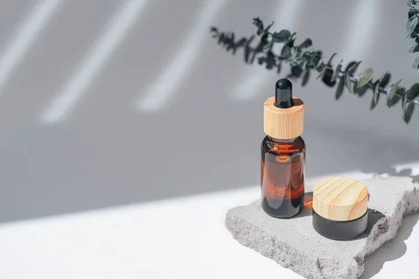 Amber bottle with dropper pipette and and jar with cream, serum or essential oil on grey concrete podium with eucalyptus branches. White background with daylight. Beauty concept for face and body care