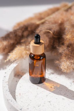 Bambu kapaklı ve kuru bitkili kehribar cam damlası. Ürün sunumu için terrazzo podyumunda bir şişe. Cilt bakım ürünleri ve kır çiçekleri. Güzellik konsepti