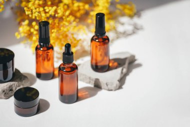 Sarı çiçekli gri beton kürsüye serumlu, tonik ya da gerekli yağlı Amber tulumba ve damlatıcı şişeler. Gündüz vakti beyaz arka plan. Yüz ve vücut bakımı için güzellik konsepti