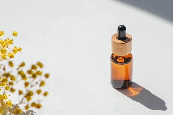 Amber bottle with cap with serum or essential oil with yellow flowers. White background with daylight and beautiful shadows. Beauty concept for face and body care