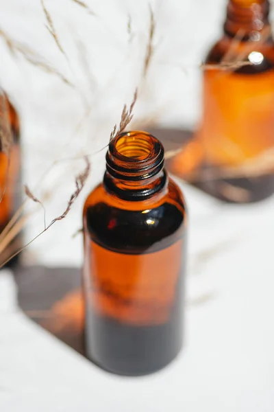 Open amber bottle with serum or essential oil with wild field grass. White background with daylight and beautiful shadows. Beauty concept for face and body care