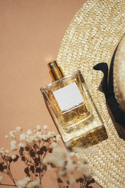 Etiketli saydam bir parfüm şişesi. Bej arka planda kumaş alışveriş çantası ve güneş şapkası. Gün ışığıyla güzel kokulu bir sunum. Doğal malzemelerde sarı tarla çiçekleriyle moda olan bir kavram. Kadın ve erkeğin özü..