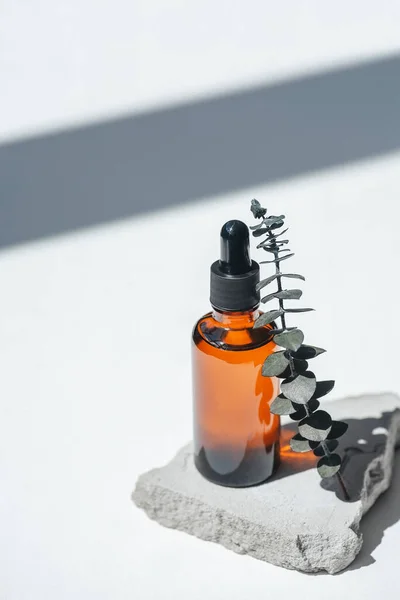 Amber bottle with dropper pipette and serum or essential oil on grey concrete podium with eucalyptus branches. White background with daylight. Beauty concept for face and body care