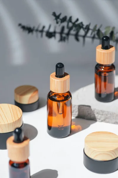 Amber bottles with dropper pipette and and jars with cream, serum or essential oil on grey concrete podium with eucalyptus branches. White background with daylight. Beauty concept for face and body care