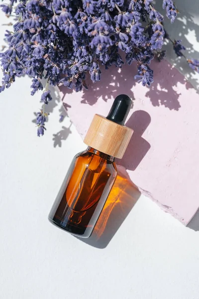 Amber dropper bottle with serum, tonic or essential oil on pink concrete podium. White background with daylight with lavender flowers. Beauty concept for face and body care