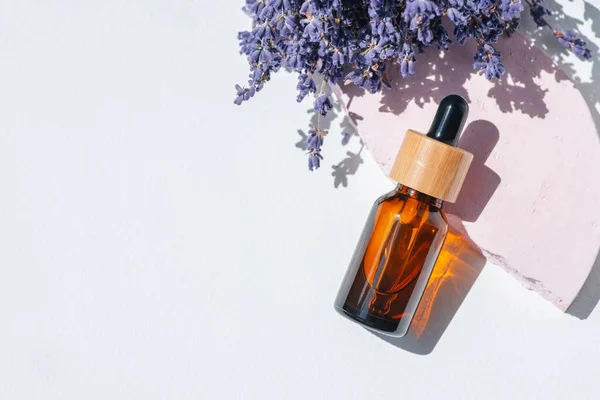 Amber dropper bottle with serum, tonic or essential oil on pink concrete podium. White background with daylight with lavender flowers. Beauty concept for face and body care