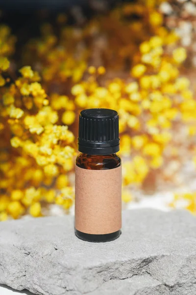 Amber bottle with craft label filled serum, tonic or essential oil on grey concrete podium with yellow flowers. White background with daylight. Beauty concept for face and body care