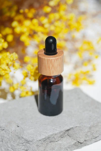 Amber dropper bottle with serum, tonic or essential oil on grey concrete podium. White background with yellow flowers.. Beauty concept for face and body care