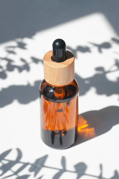 Amber bottle with dropper pipette with serum or essential oil with eucalyptus shadows. White background with daylight. Beauty concept for face and body care