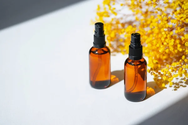 Amber pump bottles with serum, tonic or essential oil with yellow flowers. White background with daylight. Beauty concept for face and body care
