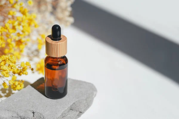 Amber dropper bottle with serum, tonic or essential oil on grey concrete podium. White background with yellow flowers.. Beauty concept for face and body care