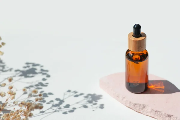 Amber bottle with cap with serum or essential oil with flowers shadows on pink platform. White background with daylight. Beauty concept for face and body care. Soothing CBD oil