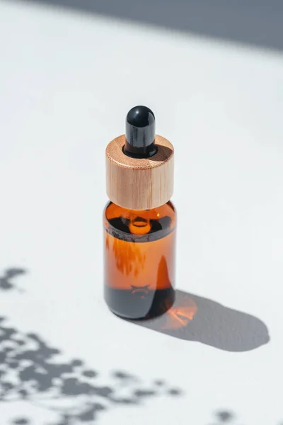 Amber bottle with cap with serum or essential oil with flowers shadows. White background with daylight. Beauty concept for face and body care. Soothing cbd oil