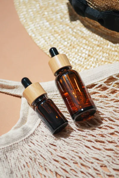 Amber bottle with dropper pipette, cloth shopping bag and sun hat on beige background. Trending concept in natural materials. Skincare serum or essential oil natural cosmetic. Beauty concept
