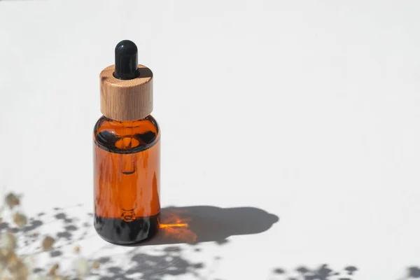 Amber bottle with cap with serum or essential oil with flowers shadows. White background with daylight. Beauty concept for face and body care. Soothing CBD oil