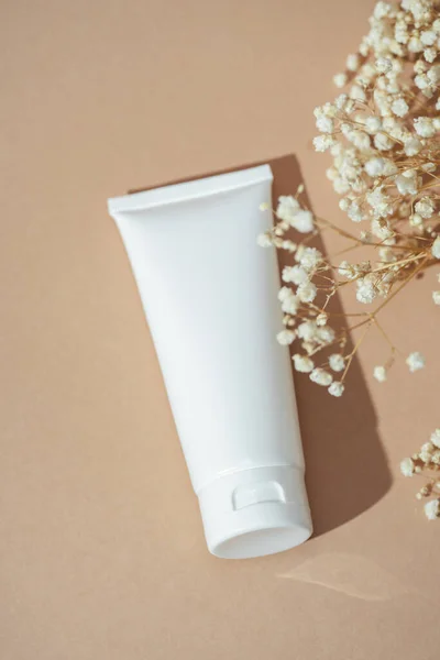 Plastic white tube for cream or lotion. Skin care or sunscreen cosmetic on beige background with beautiful flowers. Beauty concept for face care