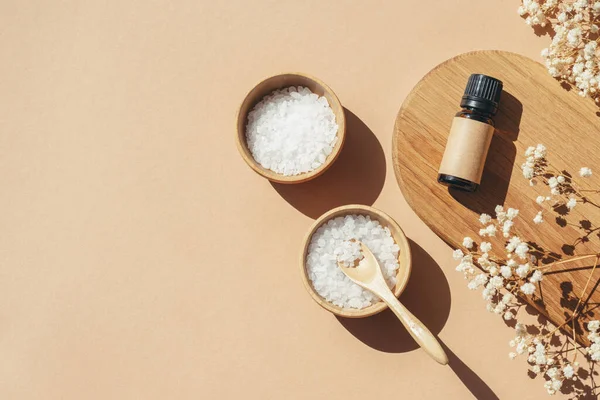 Amber bottles with craft label filled essential oil and bowls filled with white bath sea salt on wooden tray. Beige background with daylight . Beauty concept for face and body care. Copy space