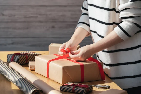 Gift wrapping in eco packaging. Young woman making bow on kraft gift box with red thread on a wooden table. Packaging and preparation of gifts for the celebration.