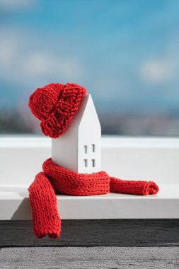Pencere pervazında kırmızı eşarp ve şapka takılı minyatür bir ev. Pasif ev ısıtma kavramı. Bir binanın ya da konutun termal yalıtımı. Enerji krizi.