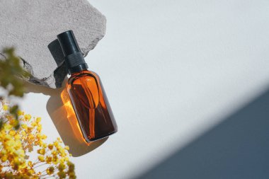 Amber pump bottle with serum, tonic or essential oil on grey concrete podium with yellow flowers. White background with daylight. Beauty concept for face and body care