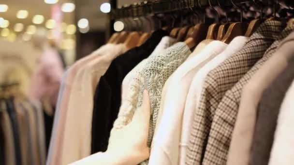 Chica elige la ropa de punto que cuelga de la percha en un primer plano tienda. — Vídeos de Stock