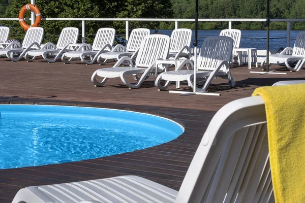Relaxing by pool concept. Yellow bath towel on back of white chaise longue, wooden podium by round pool outdoors on sunny day, hanging life preserver. Hotel spa services, healthy relax. Copy space