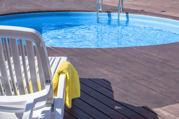Relaxing by pool concept. Yellow bath towel on armrest of white chaise longue, wooden podium by round pool outdoors on sunny day. Hotel and country club, spa services of healthy relaxation.Copy space.