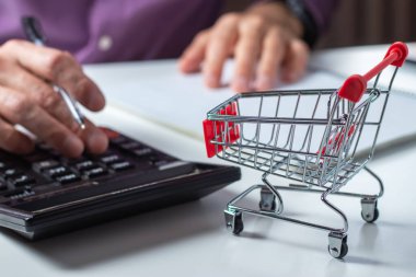 Adam bütçeyi hesaplıyor. Alışveriş arabası hesap makinesi ve kağıt ile masaya. Fakir ve düşük gelirli aile bütçesi. Artan gıda ve market fiyatları ve pahalı günlük tüketim malları kavramı