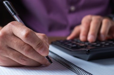 Ofiste hesap makinesi olan bir adamın elleri ve beyaz masadaki sayıları analiz eden finansal veriler..