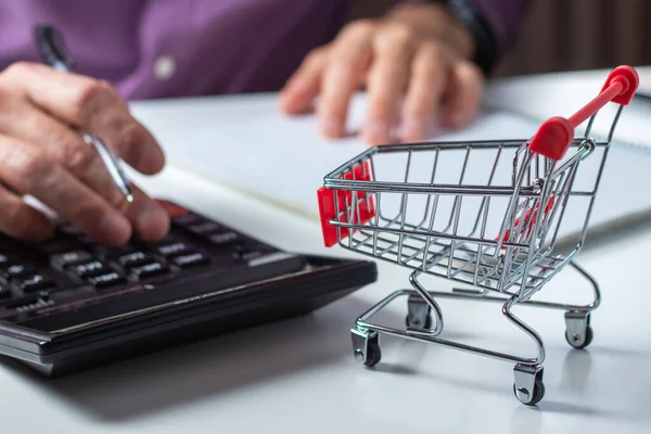 Adam bütçeyi hesaplıyor. Alışveriş arabası hesap makinesi ve kağıt ile masaya. Fakir ve düşük gelirli aile bütçesi. Artan gıda ve market fiyatları ve pahalı günlük tüketim malları kavramı