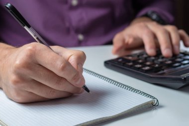 Ofiste hesap makinesi olan bir adamın elleri ve beyaz masadaki sayıları analiz eden finansal veriler..