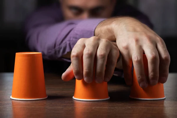 Man shows shell game of thimbles with coin, dark background. Concept deception, sleight hand