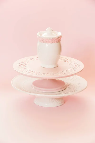 Empty white and pink cake stands and ceramic sugar pot, sugar serving bowl on top. Pink background with copy space, selective focus