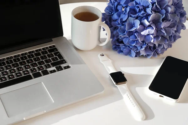 Desktop, with laptop, smart watch, phone, cup of tea and hydrangea. Business accessories on white table background. Office desktop.