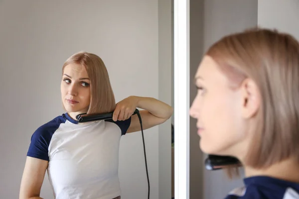 beautiful blonde straightens her hair with a hair straightener at home in front of a mirror