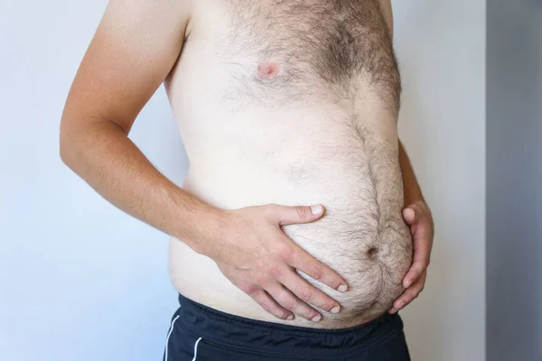 a man holds a big belly against a white wall. Obesity, beer belly