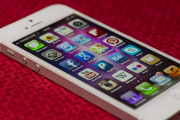 Close up of an illuminated Apple iPhone 5 screen on a red surface showing the App Store and various apps, including YouTube, Wikipedia, Google, Skype, Pinterest, Facebook, Behance, Instagram and more — Stock Photo, Image