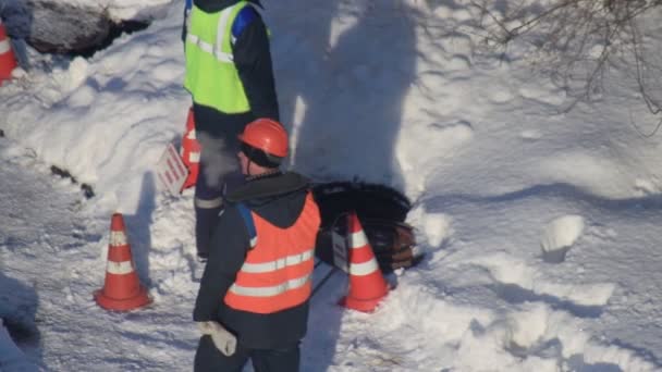 Pracownicy w kamizelkach sygnalizacyjnych wykonują prace w włazie kanalizacyjnym w celu układania komunikacji zimą, w tle — Wideo stockowe