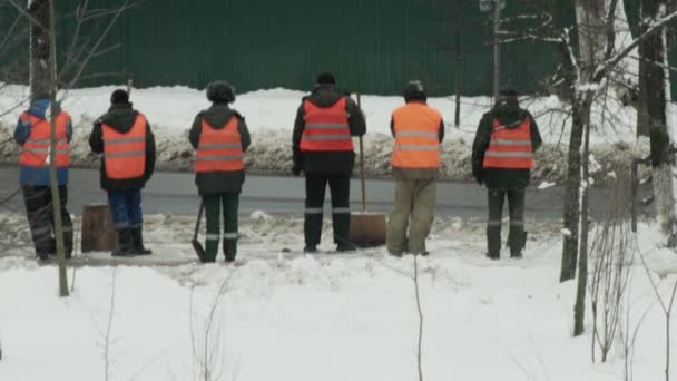 Oddział woźnych w pomarańczowych kamizelkach sygnalizacyjnych czyści śnieg w mieście na tle ciągnika do czyszczenia śniegu z dróg — Wideo stockowe
