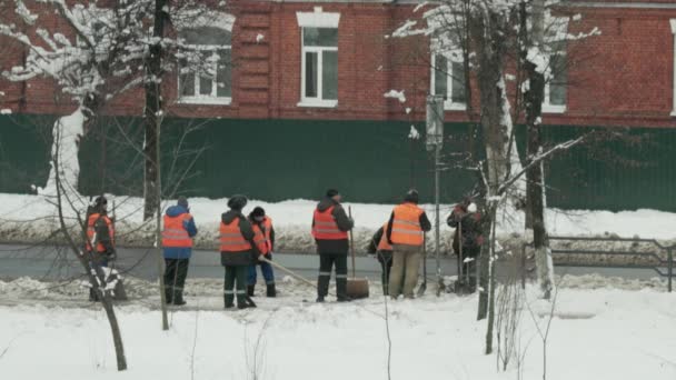 Oddział woźnych w pomarańczowych kamizelkach sygnalizacyjnych czyści śnieg w mieście na tle ciągnika do czyszczenia śniegu z dróg — Wideo stockowe